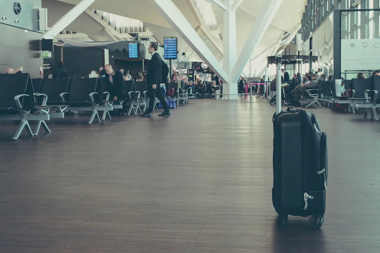 A black wheeled suitcase standing on a wooden floor in an airport departure lounge, with rows of seats and departure screens behind