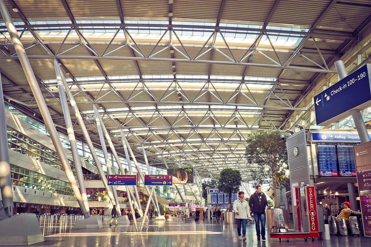 A bright airport terminal hall with a steel and glass roof, indoor trees and departure screens