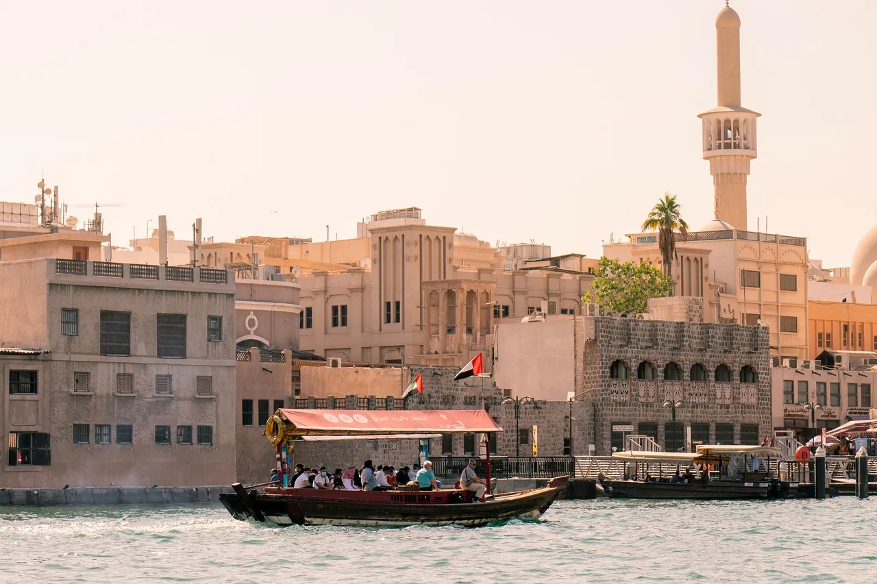 A wooden abra water taxi crossing Dubai Creek in front of old waterside buildings and a minaret at sunset