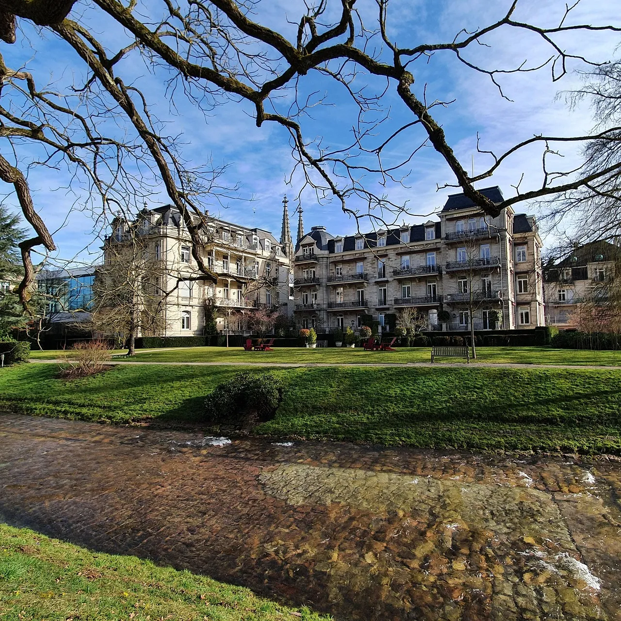 A grand white hotel in Baden-Baden behind a lawn and the little river Oos, framed by bare spring branches