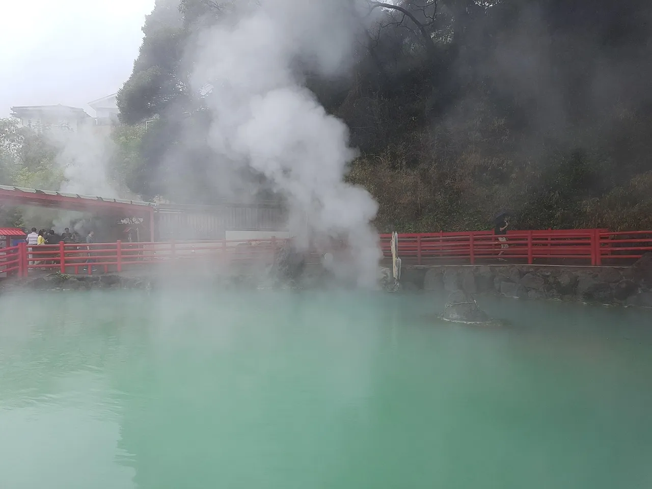 A steaming turquoise hot spring pool at one of Beppu's hells, with a red railing and green hillside behind