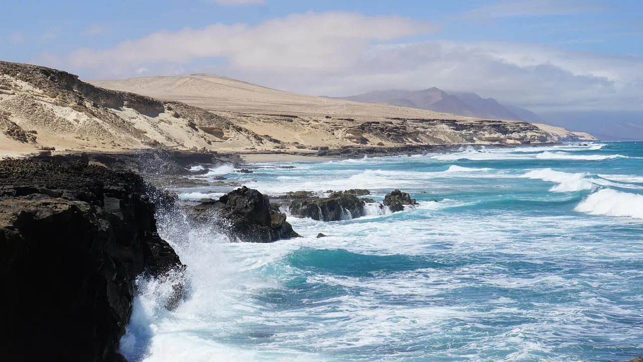 Atlantic waves breaking on dark lava rocks below sandy hills on the coast of Fuerteventura