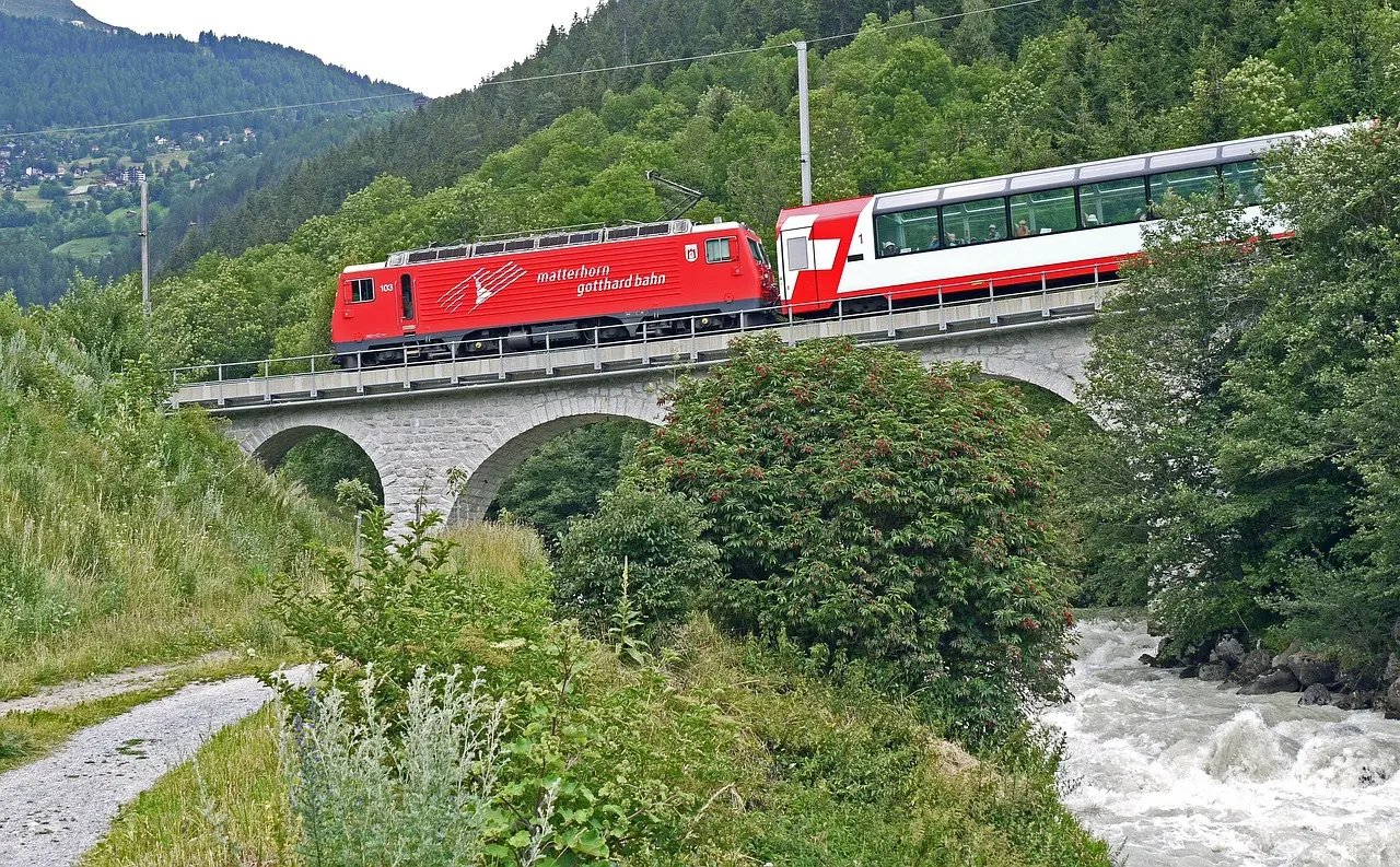 A red and white Glacier Express train crossing a stone viaduct above a rushing mountain stream in a green Swiss valley