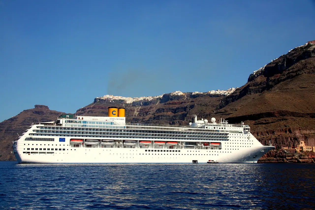 A white cruise ship anchored below the steep volcanic cliffs of Santorini, with white houses along the top
