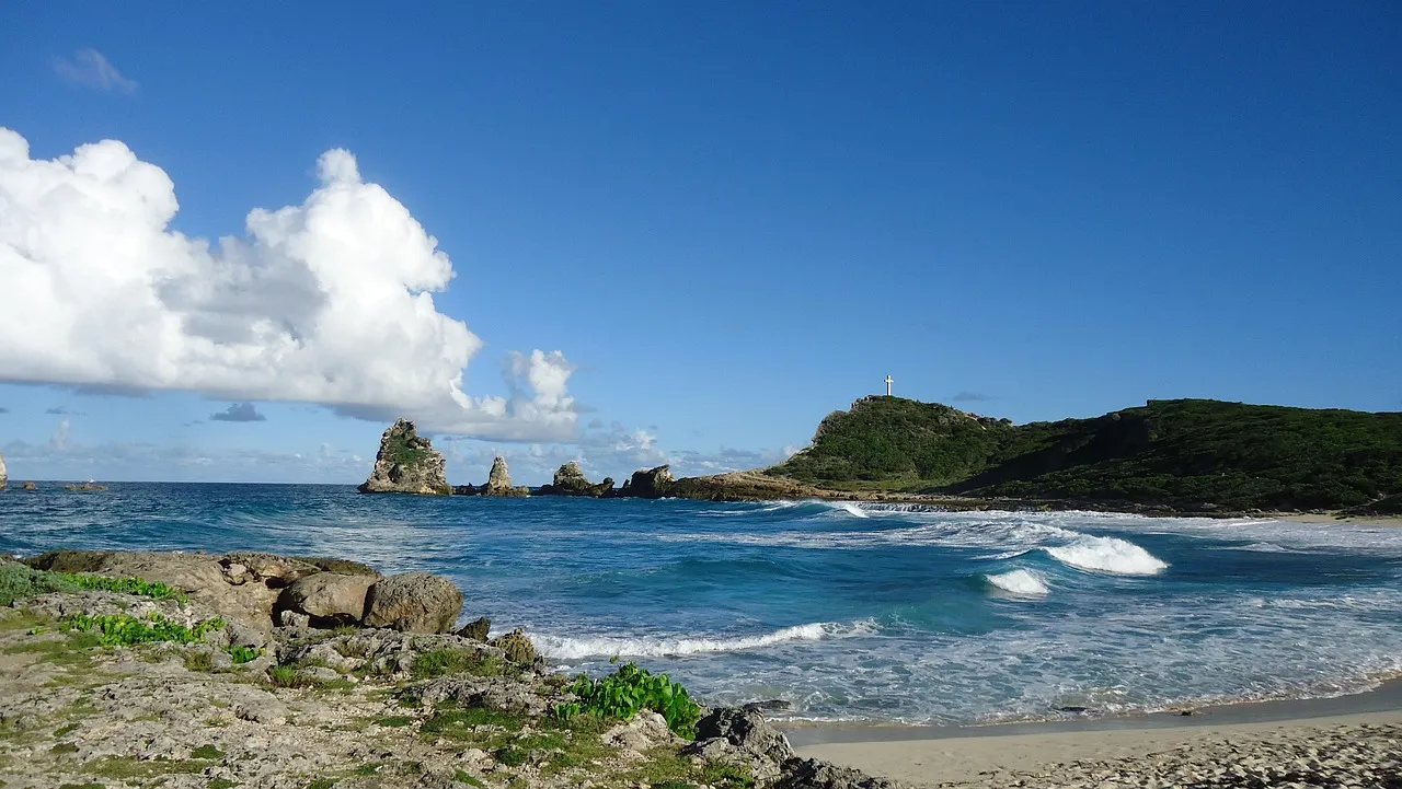 Waves rolling onto a sandy beach at Pointe des Châteaux in Guadeloupe, with rock stacks and a green headland topped by a cross
