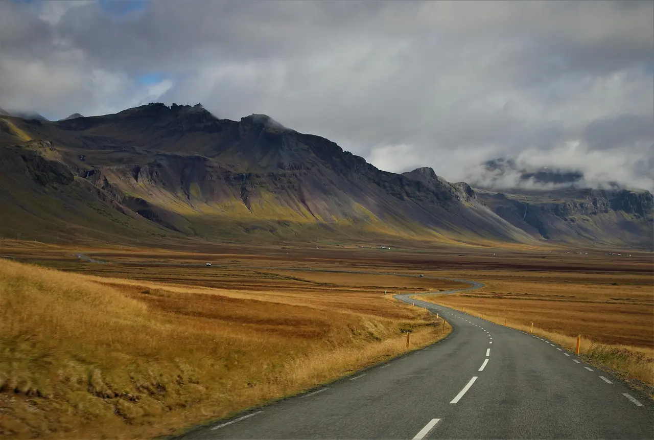 A quiet road curving through golden grassland towards dark mountains under heavy cloud in Iceland