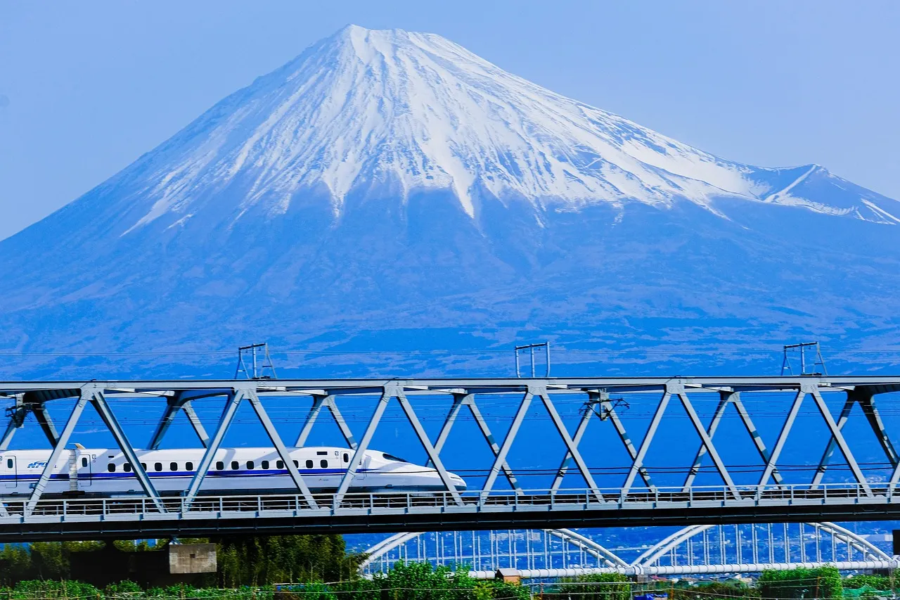 A white shinkansen bullet train crossing a bridge below the snow-capped cone of Mount Fuji under a clear blue sky