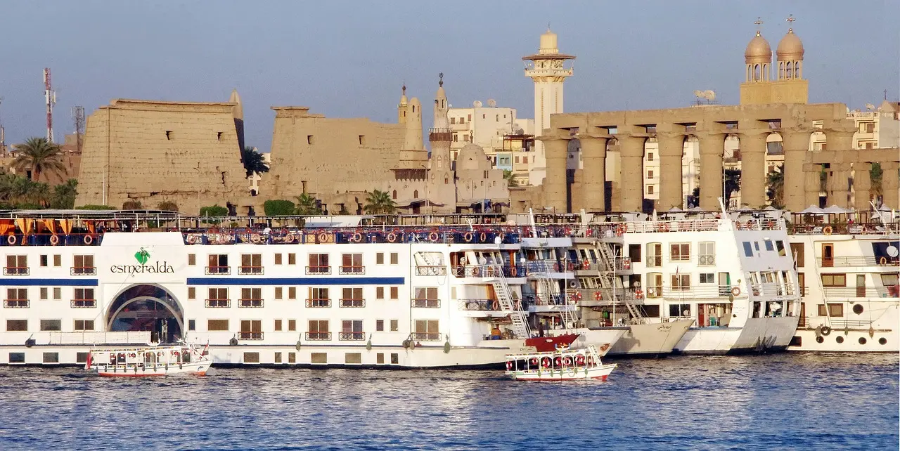 Nile cruise boats moored side by side on the river at Luxor, with the columns of Luxor Temple behind