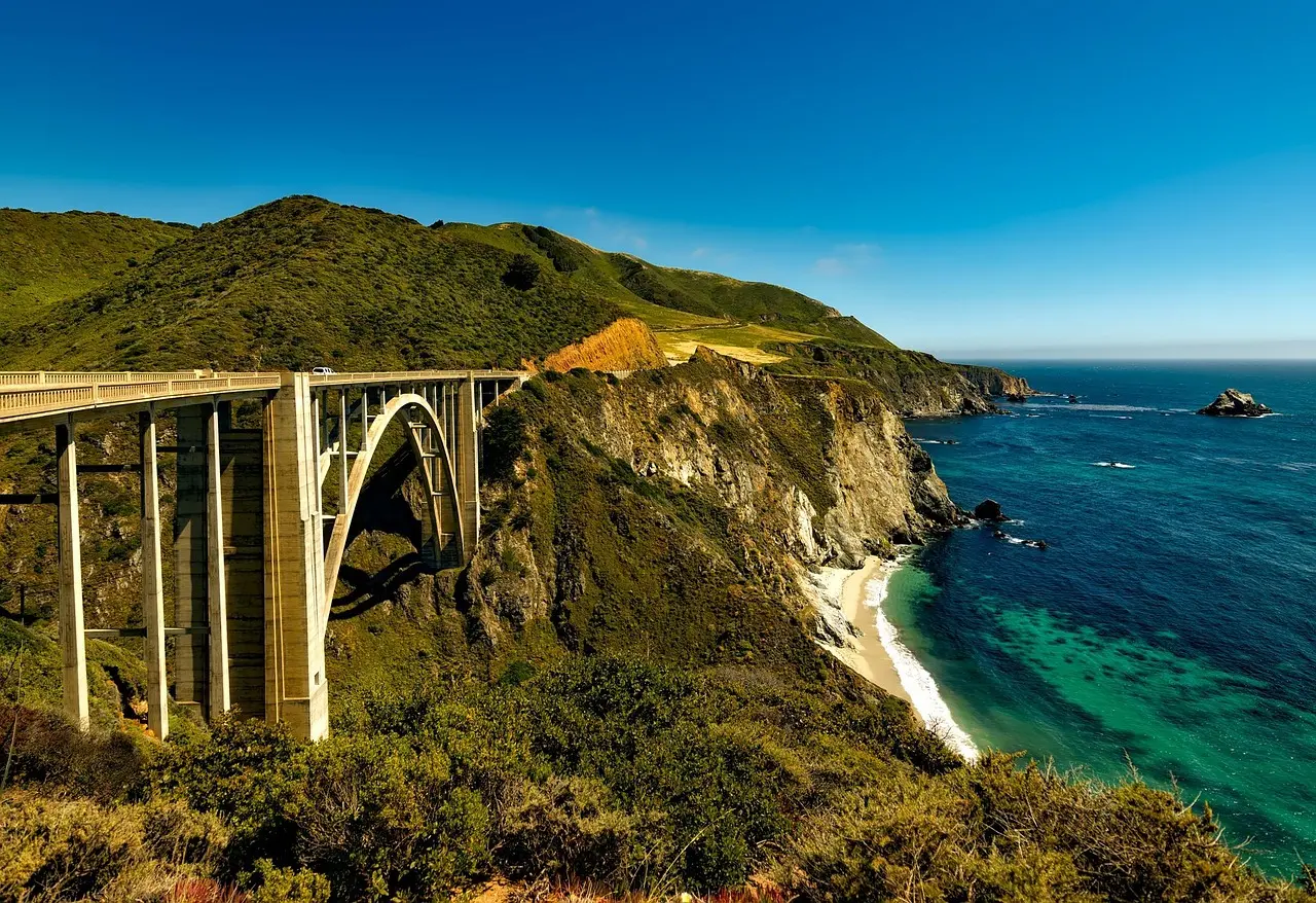 Bixby Creek Bridge arching over a green canyon on the Big Sur coast, with turquoise sea and a sandy cove below