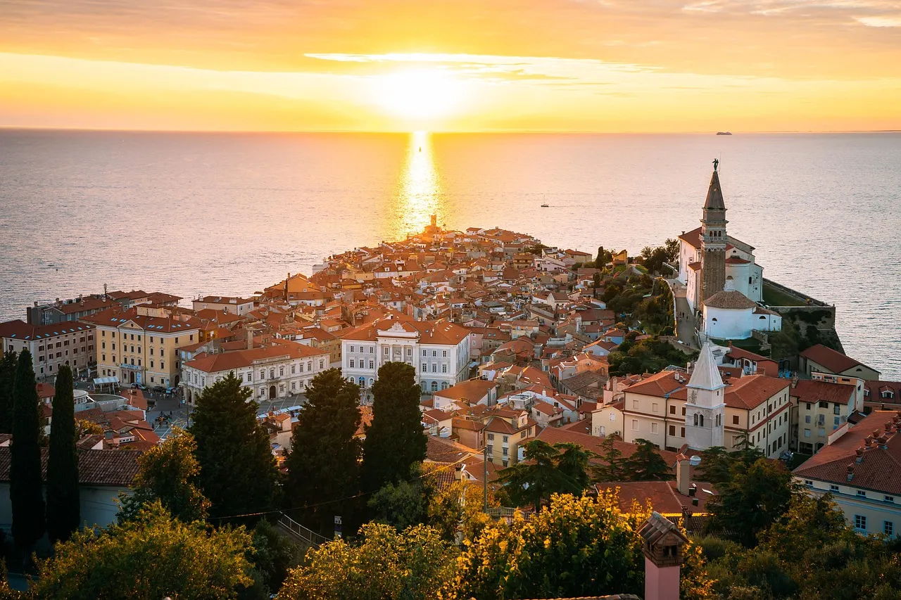 Piran's red-roofed old town and church bell tower on a headland, with the sun setting over the Adriatic Sea