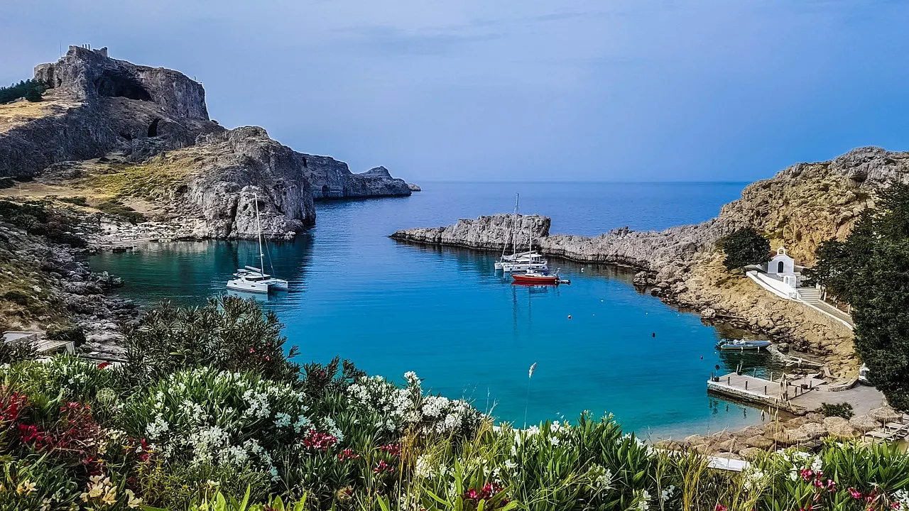 A turquoise bay with boats at anchor below a rocky headland at Lindos on Rhodes, with flowers in the foreground