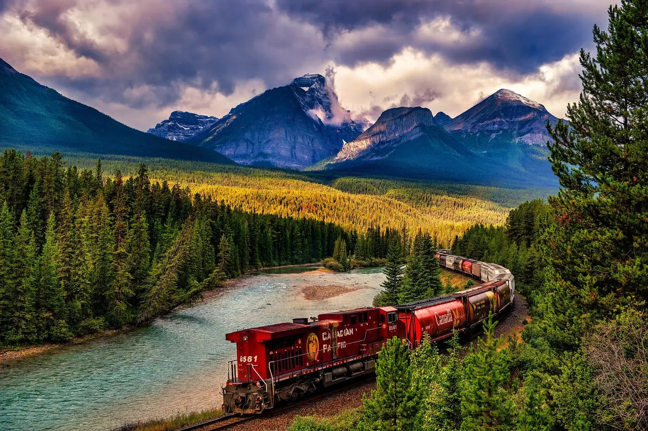 A red train curving beside the Bow River through pine forest, with jagged Rocky Mountain peaks behind