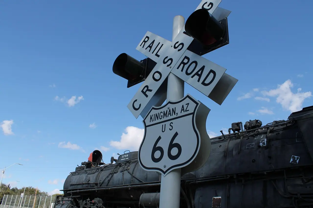 A Route 66 shield for Kingman, Arizona, below a railroad crossing sign, with an old steam locomotive behind
