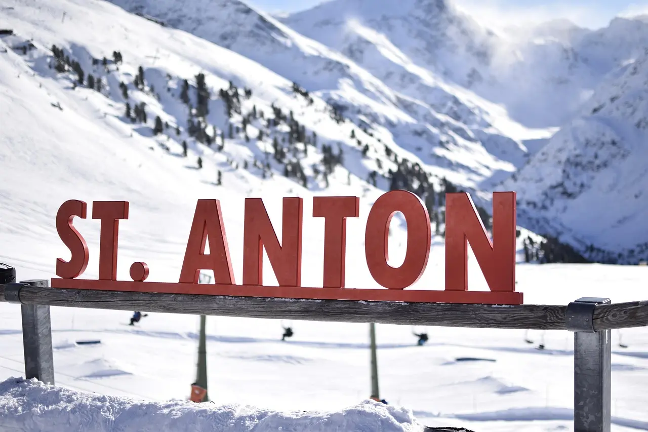 Big red letters spelling St. Anton on a snowy ridge, with skiers and snow-covered mountains behind