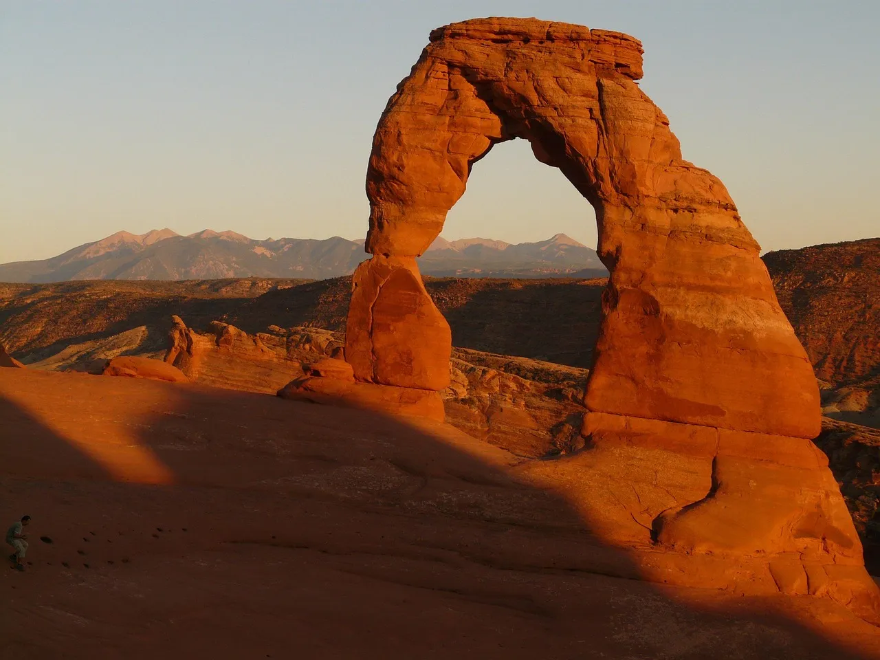 Delicate Arch in Arches National Park, Utah, glowing orange in low sunlight with snowy mountains far behind